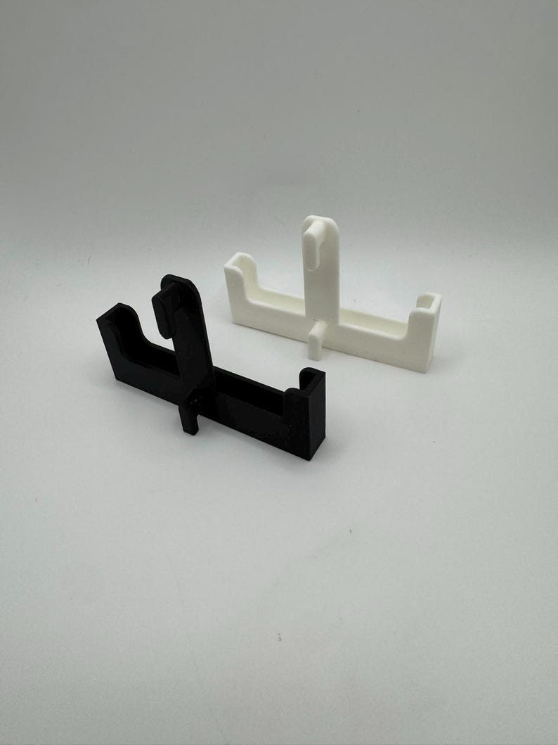 Puede incluir: Dos objetos geom&eacute;tricos de pl&aacute;stico, uno negro y otro blanco. Ambos tienen una base rectangular con una secci&oacute;n elevada y una proyecci&oacute;n en forma de gancho. Los objetos est&aacute;n dise&ntilde;ados para sujetar o soportar algo.