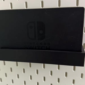 May include: A black Nintendo Switch console holder with a pegboard background. The holder has a small shelf for the console and a larger shelf for the Joy-Cons. The console is visible in the holder, with the Nintendo Switch logo and text visible.