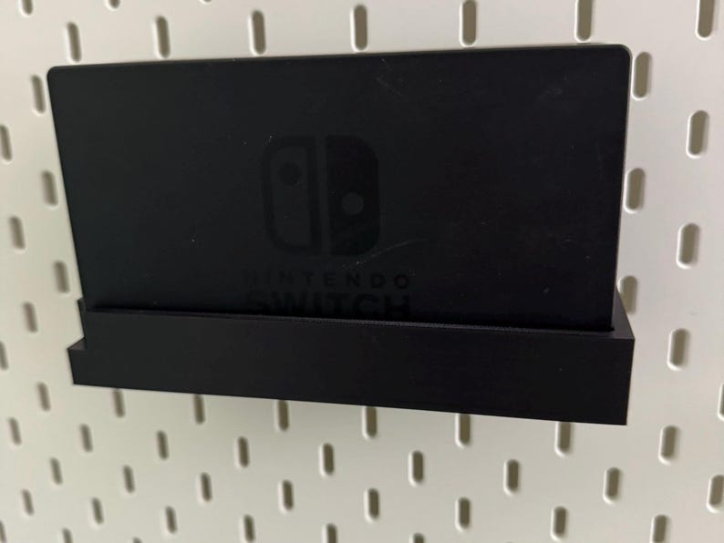 May include: A black Nintendo Switch console holder with a pegboard background. The holder has a small shelf for the console and a larger shelf for the Joy-Cons. The console is visible in the holder, with the Nintendo Switch logo and text visible.