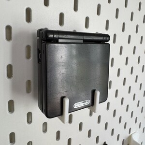 May include: A black Nintendo Game Boy Advance SP handheld console mounted on a white pegboard. The console is held in place by two white brackets. The Nintendo logo is visible on the console.
