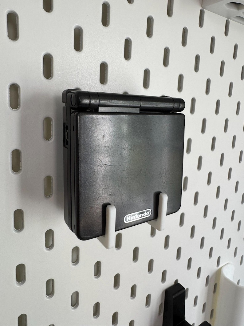 May include: A black Nintendo Game Boy Advance SP handheld console mounted on a white pegboard. The console is held in place by two white brackets. The Nintendo logo is visible on the console.