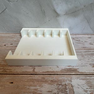 May include: A white, rectangular organizer tray with multiple slots and dividers. The tray is made of a solid, opaque material and sits on a wooden surface. The design includes vertical dividers and a raised edge.