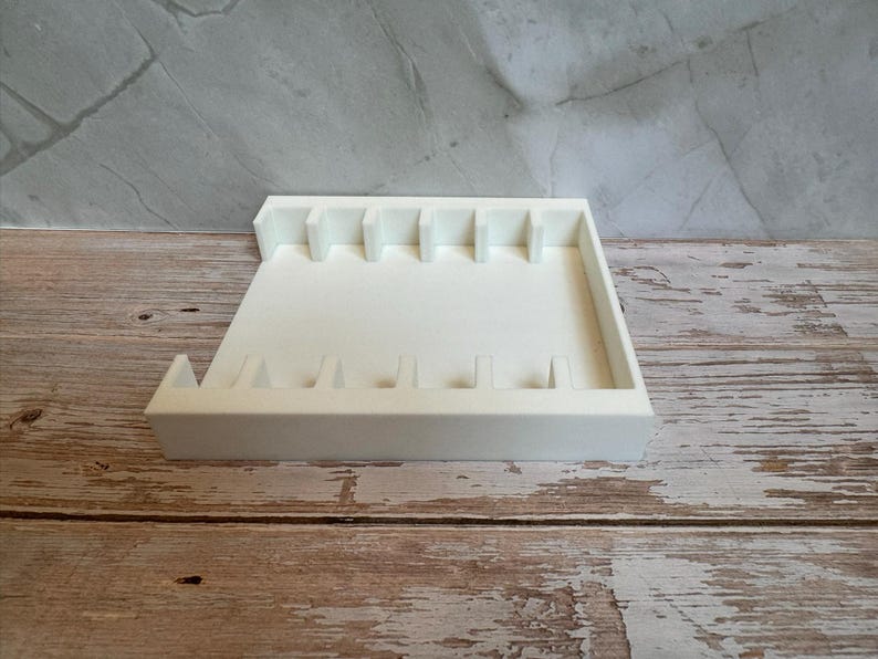 May include: A white, rectangular organizer tray with multiple slots and dividers. The tray is made of a solid, opaque material and sits on a wooden surface. The design includes vertical dividers and a raised edge.