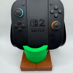 May include: Black Nintendo Switch console with blue and orange controllers, on a green and brown stand. The console has the Nintendo Switch logo and button controls. The stand is brown and has a green support.