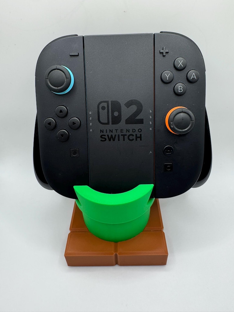 May include: Black Nintendo Switch console with blue and orange controllers, on a green and brown stand. The console has the Nintendo Switch logo and button controls. The stand is brown and has a green support.