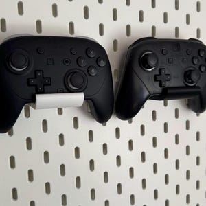 K&ouml;nnte beinhalten: Zwei schwarze Gamecontroller sind an einer wei&szlig;en Lochwand befestigt. Die Controller haben verschiedene Tasten, Joysticks und Richtungspads. Jeder Controller wird von einem wei&szlig;en Halter gehalten. Die Controller sind f&uuml;r eine Spielekonsole.