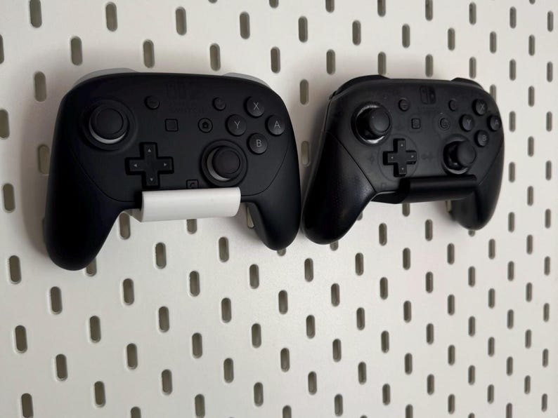 K&ouml;nnte beinhalten: Zwei schwarze Gamecontroller sind an einer wei&szlig;en Lochwand befestigt. Die Controller haben verschiedene Tasten, Joysticks und Richtungspads. Jeder Controller wird von einem wei&szlig;en Halter gehalten. Die Controller sind f&uuml;r eine Spielekonsole.