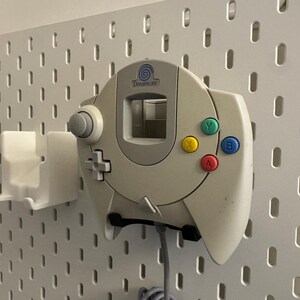 K&ouml;nnte beinhalten: Cremefarbener Dreamcast-Controller mit farbigen Kn&ouml;pfen (gr&uuml;n, gelb, blau, rot) an einer wei&szlig;en Lochwand befestigt. Der Controller ist an einer wei&szlig;en Halterung befestigt. Der Controller hat einen grauen Joystick und ein Steuerkreuz.