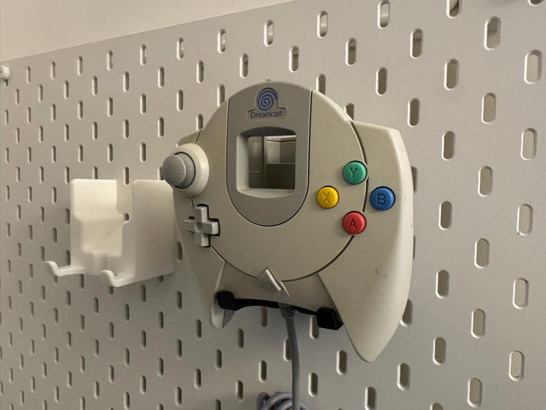 K&ouml;nnte beinhalten: Cremefarbener Dreamcast-Controller mit farbigen Kn&ouml;pfen (gr&uuml;n, gelb, blau, rot) an einer wei&szlig;en Lochwand befestigt. Der Controller ist an einer wei&szlig;en Halterung befestigt. Der Controller hat einen grauen Joystick und ein Steuerkreuz.
