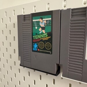 Pode incluir: Cartucho de jogo cinza do Nintendo Entertainment System para "Tennis" com um r&oacute;tulo verde e preto com um jogador de t&ecirc;nis. O cartucho &eacute; montado em uma placa perfurada branca.