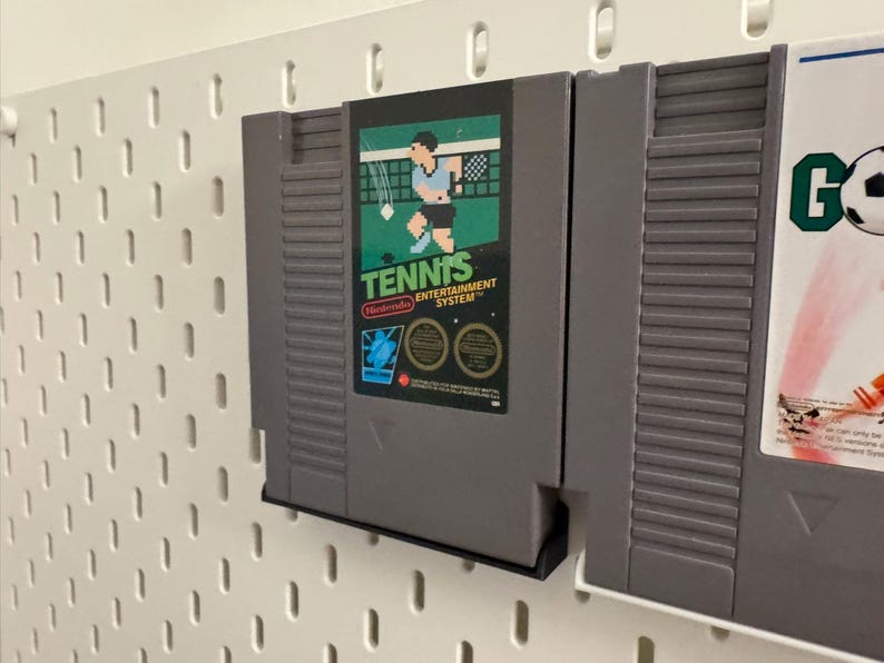 Pode incluir: Cartucho de jogo cinza do Nintendo Entertainment System para "Tennis" com um r&oacute;tulo verde e preto com um jogador de t&ecirc;nis. O cartucho &eacute; montado em uma placa perfurada branca.