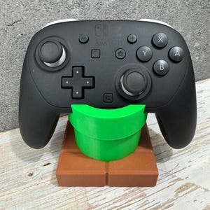 K&ouml;nnte beinhalten: Ein schwarzer Nintendo Switch Pro Controller ruht auf einem braunen und gr&uuml;nen St&auml;nder. Der St&auml;nder ist so gestaltet, dass er wie ein gr&uuml;nes Rohr auf einer braunen Ziegelplattform aussieht. Der Controller hat wei&szlig;e Akzente und verschiedene Tasten.