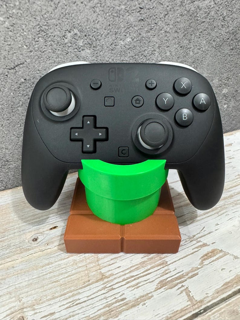 K&ouml;nnte beinhalten: Ein schwarzer Nintendo Switch Pro Controller ruht auf einem braunen und gr&uuml;nen St&auml;nder. Der St&auml;nder ist so gestaltet, dass er wie ein gr&uuml;nes Rohr auf einer braunen Ziegelplattform aussieht. Der Controller hat wei&szlig;e Akzente und verschiedene Tasten.
