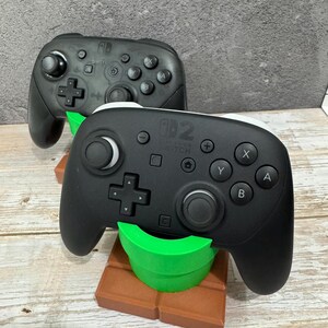 K&ouml;nnte beinhalten: Zwei schwarze Nintendo Switch Pro Controller. Ein Controller ruht auf einem braunen und gr&uuml;nen St&auml;nder in Form eines Videospielrohrs. Die Controller haben Tasten mit den Bezeichnungen A, B, X und Y.