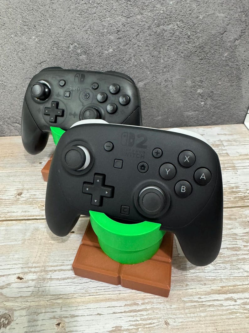 K&ouml;nnte beinhalten: Zwei schwarze Nintendo Switch Pro Controller. Ein Controller ruht auf einem braunen und gr&uuml;nen St&auml;nder in Form eines Videospielrohrs. Die Controller haben Tasten mit den Bezeichnungen A, B, X und Y.