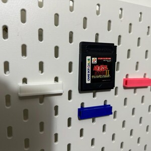 Pode incluir: Um quadro de pinos branco com um cartucho de jogo Game Boy Color preto, uma prateleira de pl&aacute;stico branca, uma prateleira de pl&aacute;stico rosa e uma prateleira de pl&aacute;stico azul.