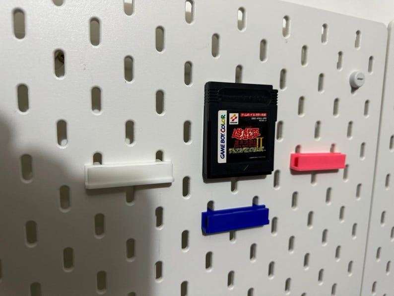Pode incluir: Um quadro de pinos branco com um cartucho de jogo Game Boy Color preto, uma prateleira de pl&aacute;stico branca, uma prateleira de pl&aacute;stico rosa e uma prateleira de pl&aacute;stico azul.