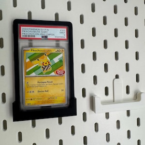 Peut inclure: Carte Pokémon Pikachu encadrée, affichée sur un panneau perforé blanc. La carte est dans un cadre noir, avec du texte visible en haut. Un crochet de panneau perforé blanc est également visible.