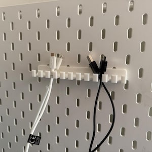 May include: A white plastic cable organizer with multiple slots, holding white and black charging cables. The organizer is mounted on a white pegboard with evenly spaced oval holes. The cables have USB and other connectors.