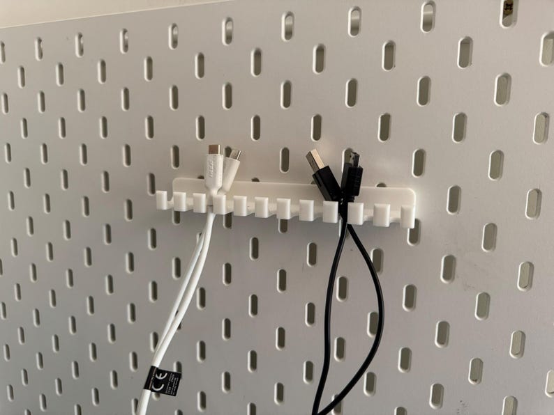 May include: A white plastic cable organizer with multiple slots, holding white and black charging cables. The organizer is mounted on a white pegboard with evenly spaced oval holes. The cables have USB and other connectors.