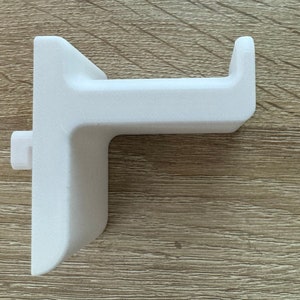 Pu&ograve; includere: Un supporto per cuffie bianco, stampato in 3D. Il supporto ha una base rettangolare con un braccio a forma di gancio che si estende verso l'esterno. Il design include un piccolo perno rettangolare sul lato per un montaggio sicuro. L'oggetto &egrave; in materiale bianco.