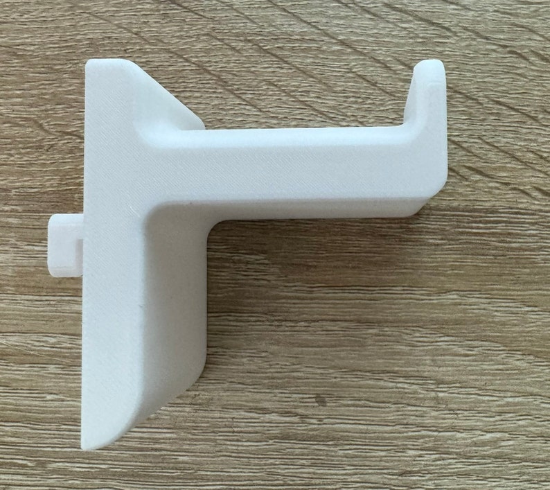 Pu&ograve; includere: Un supporto per cuffie bianco, stampato in 3D. Il supporto ha una base rettangolare con un braccio a forma di gancio che si estende verso l'esterno. Il design include un piccolo perno rettangolare sul lato per un montaggio sicuro. L'oggetto &egrave; in materiale bianco.