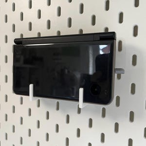 K&ouml;nnte beinhalten: Eine schwarze Handheld-Spielkonsole, die mit wei&szlig;en und grauen Halterungen an einer wei&szlig;en Lochwand befestigt ist. Die Konsole ist rechteckig mit gl&auml;nzendem Finish und sichtbarem Kameraobjektiv. Die Lochwand hat gleichm&auml;&szlig;ig verteilte ovale L&ouml;cher.