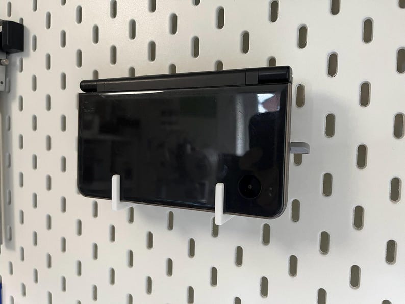 K&ouml;nnte beinhalten: Eine schwarze Handheld-Spielkonsole, die mit wei&szlig;en und grauen Halterungen an einer wei&szlig;en Lochwand befestigt ist. Die Konsole ist rechteckig mit gl&auml;nzendem Finish und sichtbarem Kameraobjektiv. Die Lochwand hat gleichm&auml;&szlig;ig verteilte ovale L&ouml;cher.