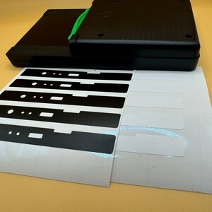 May include: A sheet of white sticker paper with six black rectangular stickers. The stickers have cutouts in the shape of a rectangle and two circles.