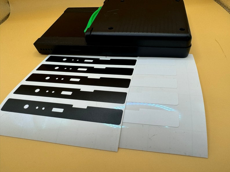 May include: A sheet of white sticker paper with six black rectangular stickers. The stickers have cutouts in the shape of a rectangle and two circles.