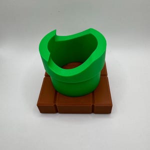 K&ouml;nnte beinhalten: Ein gr&uuml;nes Rohr, das an ein Videospiel erinnert, steht auf einer braunen, ziegelartigen Basis. Das Rohr hat eine gebogene &Ouml;ffnung und eine glatte Textur. Die Basis ist rechteckig mit horizontalen Linien, die ein Ziegelmuster bilden. Das Design hat eine spielerische, skurrile &Auml;sthetik.