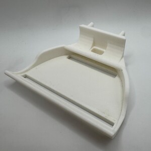 May include: A white, 3D-printed object with a curved tray and a raised back section. The tray has a rectangular opening and a raised edge. The back section has two small, cylindrical protrusions. The object is made of a solid, opaque white material.