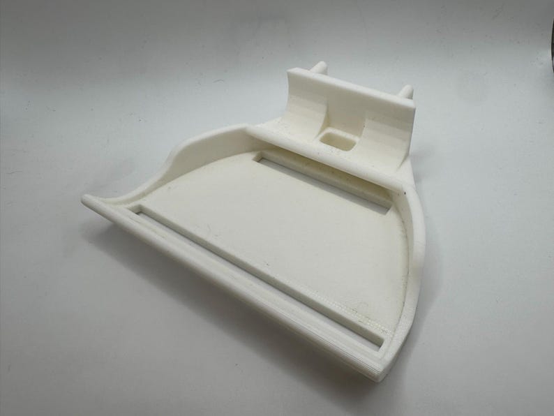 May include: A white, 3D-printed object with a curved tray and a raised back section. The tray has a rectangular opening and a raised edge. The back section has two small, cylindrical protrusions. The object is made of a solid, opaque white material.