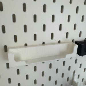 May include: A white, rectangular shelf with raised edges, designed to attach to a pegboard. The shelf is made of a smooth, solid material and is shown against a white pegboard background with evenly spaced oval holes.