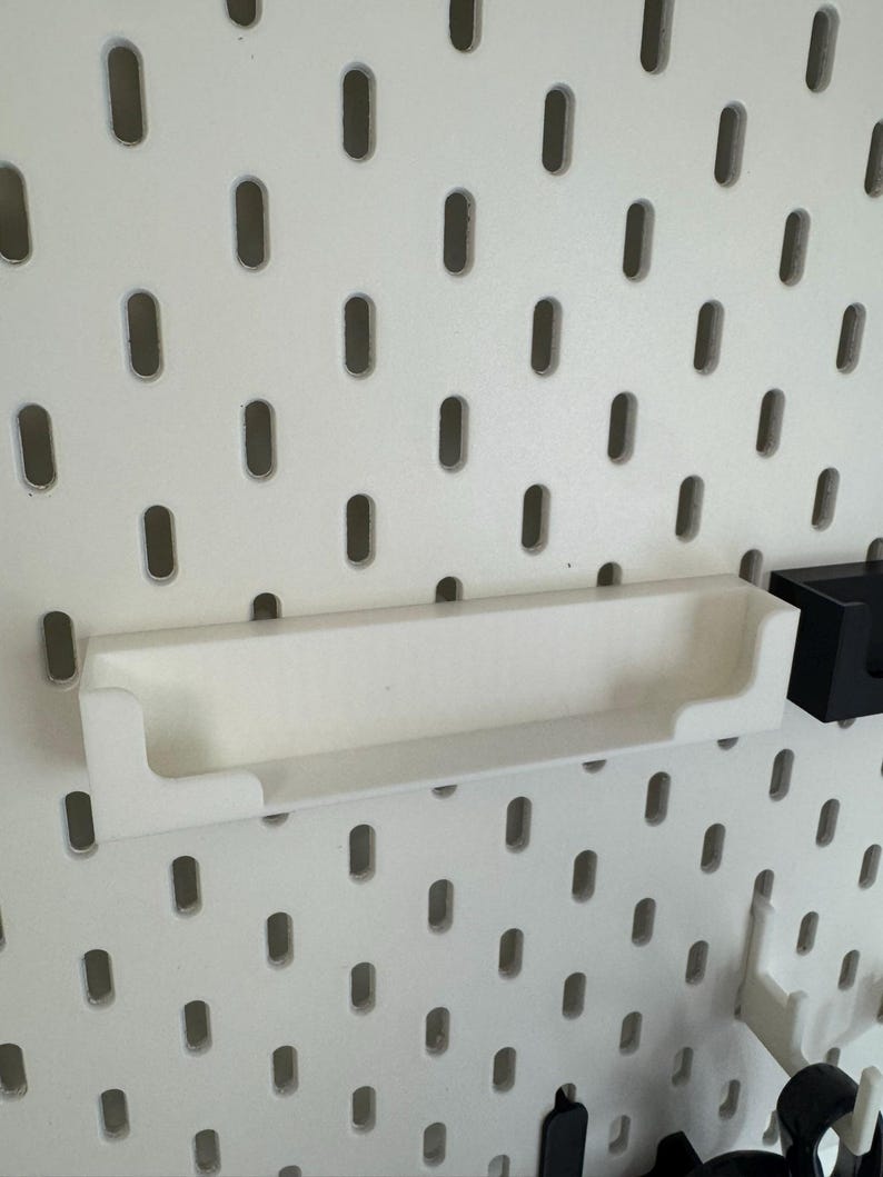 May include: A white, rectangular shelf with raised edges, designed to attach to a pegboard. The shelf is made of a smooth, solid material and is shown against a white pegboard background with evenly spaced oval holes.