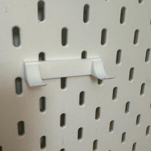 Op de afbeelding: Witte plastic pegboard haak met twee tanden. De haak is gemonteerd op een wit pegboard.