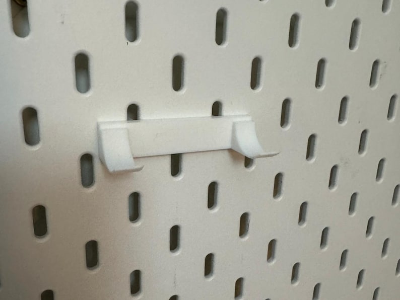 Op de afbeelding: Witte plastic pegboard haak met twee tanden. De haak is gemonteerd op een wit pegboard.