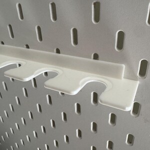 May include: A white pegboard with a white plastic shelf attachment. The shelf has three curved cutouts for holding items. The pegboard has many evenly spaced, oblong holes.