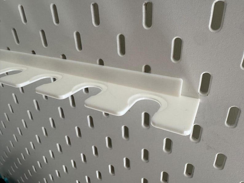 May include: A white pegboard with a white plastic shelf attachment. The shelf has three curved cutouts for holding items. The pegboard has many evenly spaced, oblong holes.