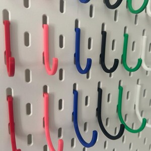 Puede incluir: Un tablero de clavijas con una variedad de ganchos en diferentes colores, incluyendo rojo, rosa, azul, negro, verde y blanco. Los ganchos tienen todos la forma de una J y est&aacute;n dispuestos en filas en el tablero de clavijas.