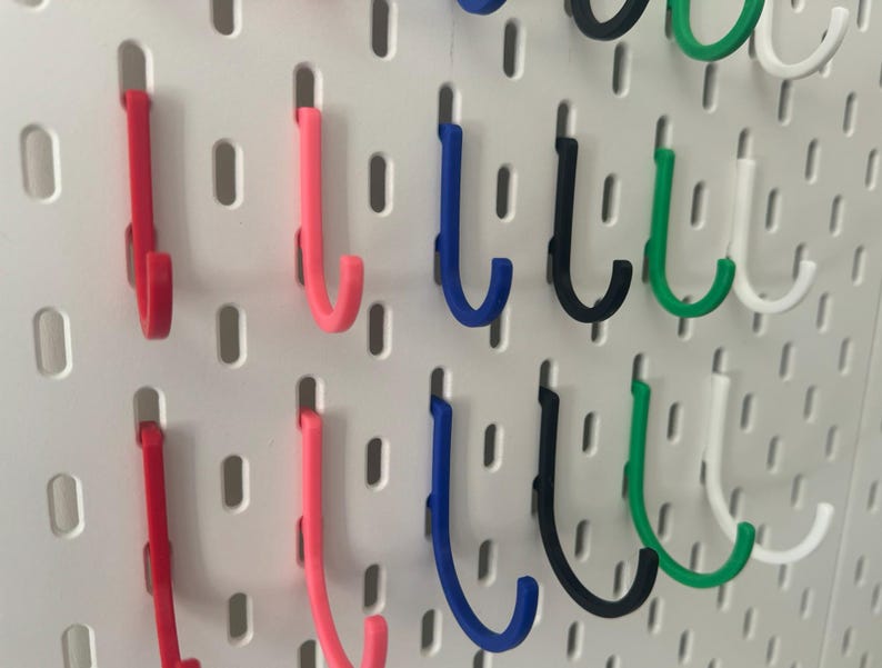 Puede incluir: Un tablero de clavijas con una variedad de ganchos en diferentes colores, incluyendo rojo, rosa, azul, negro, verde y blanco. Los ganchos tienen todos la forma de una J y est&aacute;n dispuestos en filas en el tablero de clavijas.