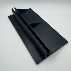 Puede incluir: Un soporte de pl&aacute;stico negro con dos ranuras para sujetar objetos. El soporte est&aacute; inclinado hacia arriba y tiene una superficie texturizada.
