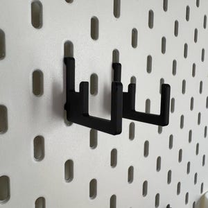May include: Two black metal pegboard hooks are mounted on a white pegboard. The hooks are designed to hold items and are made of a sturdy material. The pegboard has evenly spaced oval holes.