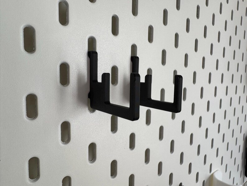 May include: Two black metal pegboard hooks are mounted on a white pegboard. The hooks are designed to hold items and are made of a sturdy material. The pegboard has evenly spaced oval holes.