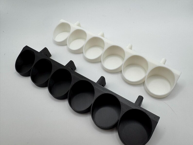 以下が含まれることがあります： 黒と白の2つの3Dプリントホルダー。それぞれに5つの円形のカップのようなコンパートメントがあります。ホルダーは平らに置くように設計されており、側面に小さなハンドル状の延長部があります。これらのオブジェクトは、小さなアイテムを整理するためのものです。
