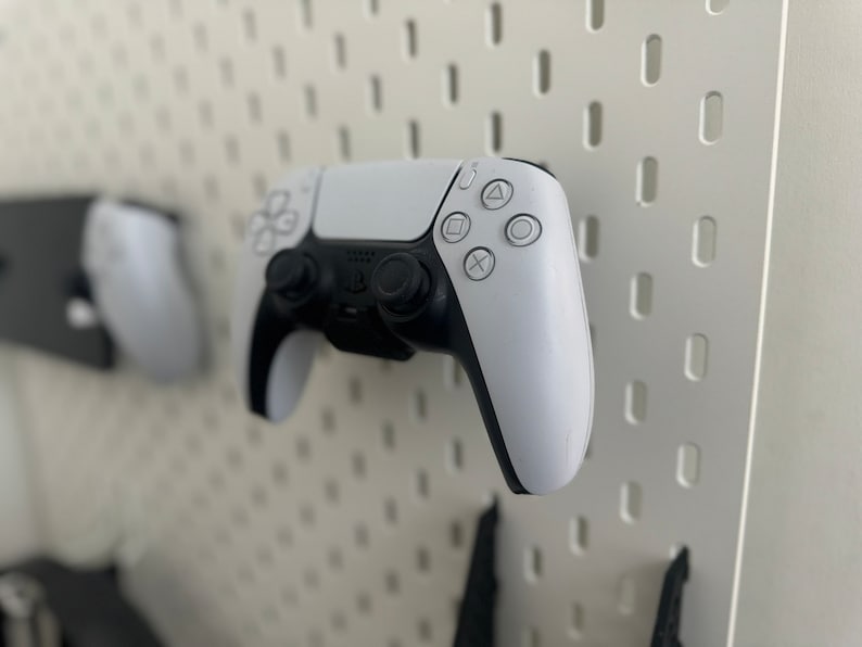 May include: A white wireless video game controller with black buttons and a black analog stick. The controller is mounted on a white pegboard.
