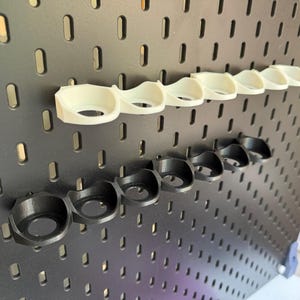 以下が含まれることがあります： 黒と白のプラスチック製のカップホルダーが2列あるペグボード。カップホルダーは、筆や工具などの小さなアイテムを収納するために設計されています。