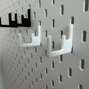 以下が含まれることがあります： 白いペグボードにフックがいくつか付いたクローズアップ写真。白いフック2つと黒いフック1つがボードに取り付けられています。フックは物を保持するように設計されており、丈夫な素材で作られています。