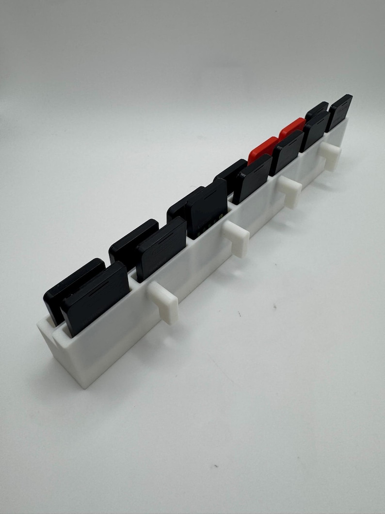 K&ouml;nnte beinhalten: Ein wei&szlig;er, 3D-gedruckter Spielkassettenhalter mit mehreren Schlitzen. Der Halter enth&auml;lt mehrere schwarze Spielkassetten und einige rote. Das Design weist kleine, wei&szlig;e St&uuml;tzstrukturen entlang der Vorderkante auf.