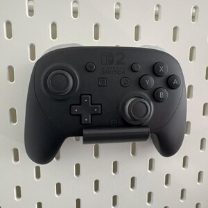K&ouml;nnte beinhalten: Ein schwarzer Nintendo Switch Pro Controller mit grauen Akzenten ist an einer wei&szlig;en Lochwand befestigt. Der Controller verf&uuml;gt &uuml;ber ein Steuerkreuz, Tasten mit den Bezeichnungen X, Y, A und B sowie zwei Analog-Sticks. Das Nintendo Switch-Logo ist sichtbar.